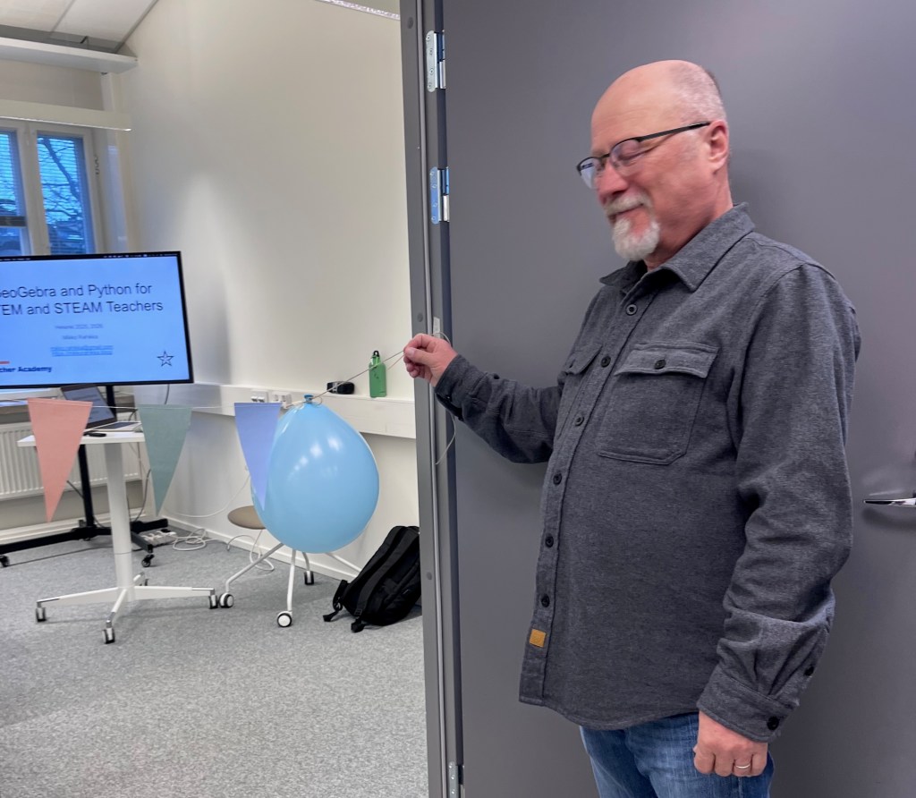 Mies pitelee sinistä ilmapalloa oven edessä luokkahuoneessa, taustalla esitysnäyttö ja koristeita.