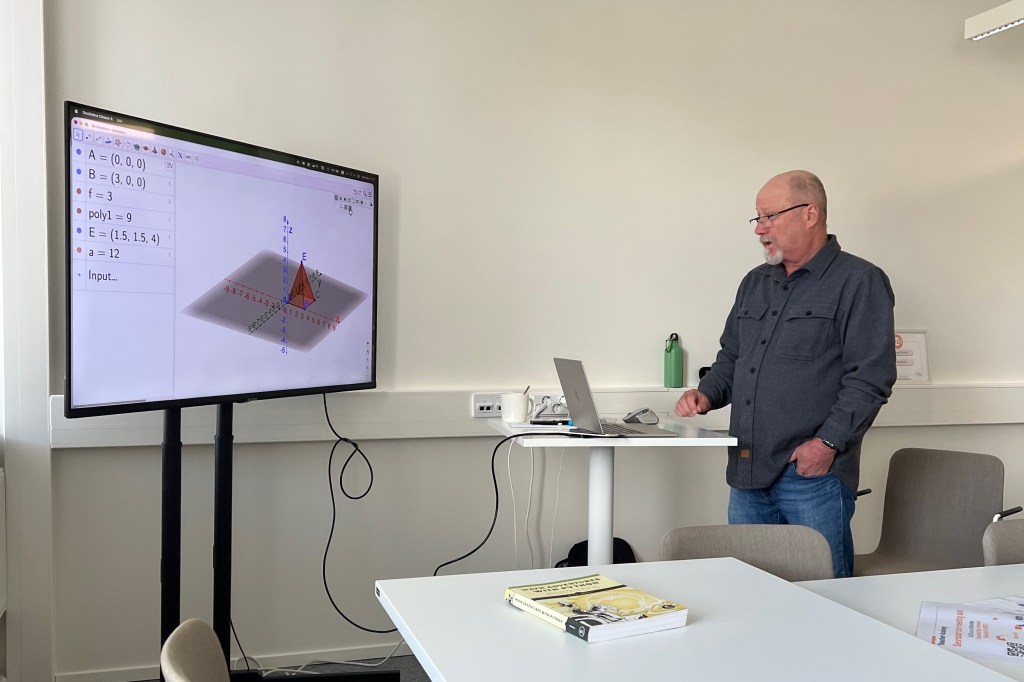 Mies seisoo tietokoneen vieressä ja esittelee 3D-mallia suurilla näytöllä. Näytöllä näkyy geometrisiä kaavioita ja laskentatietoja.